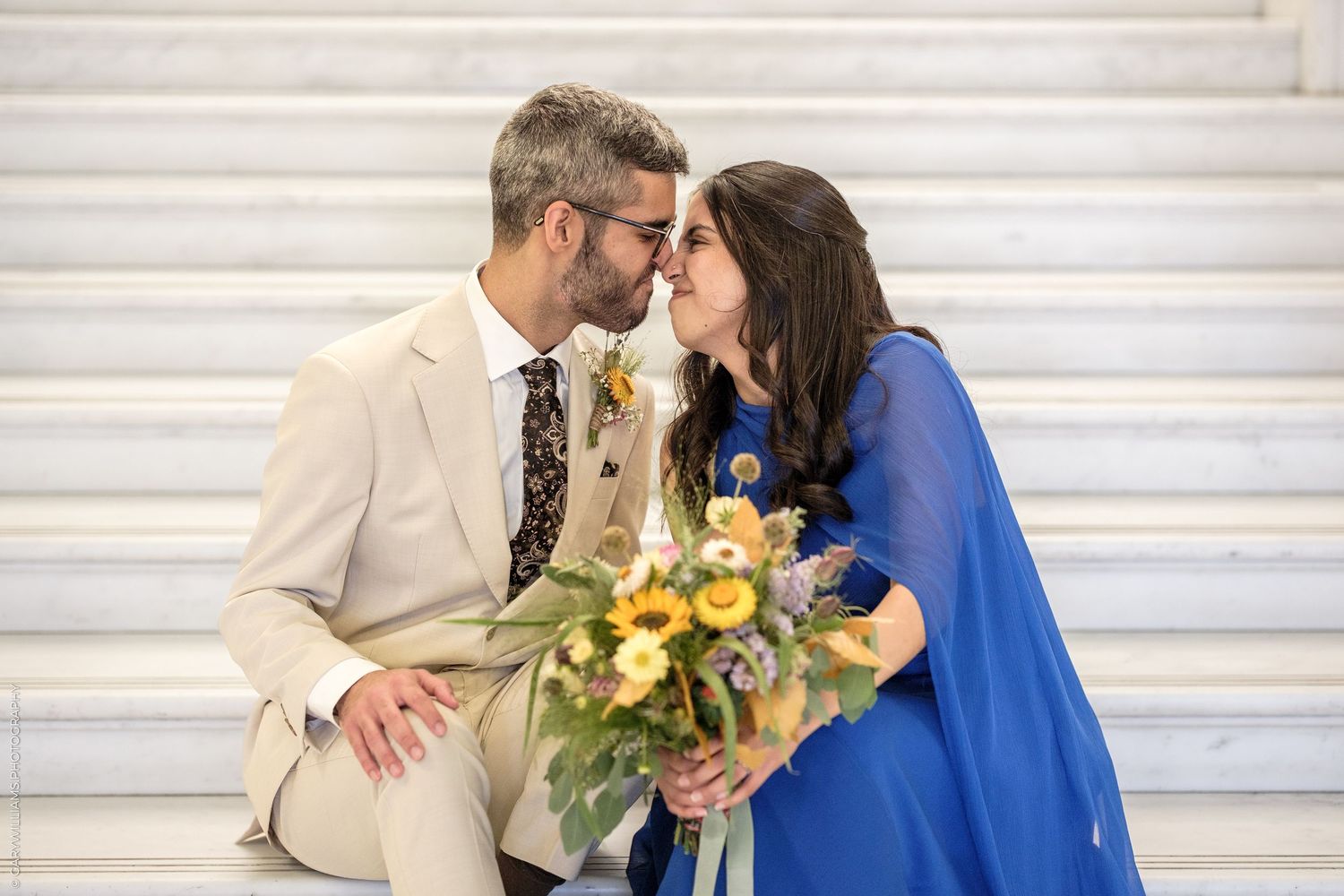 Couple in wedding attire sharing a romantic moment on marble steps, holding a yellow and blue floral bouquet.