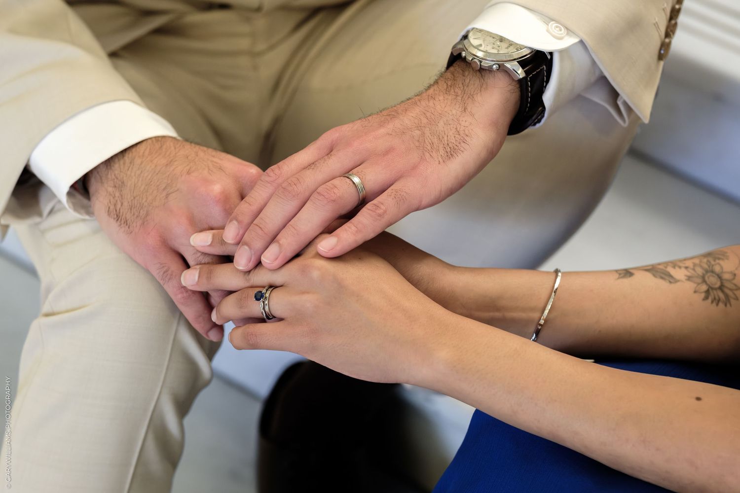 Close-up of a couple holding hands showing wedding rings, mans watch, and womans bracelet and tattoo.