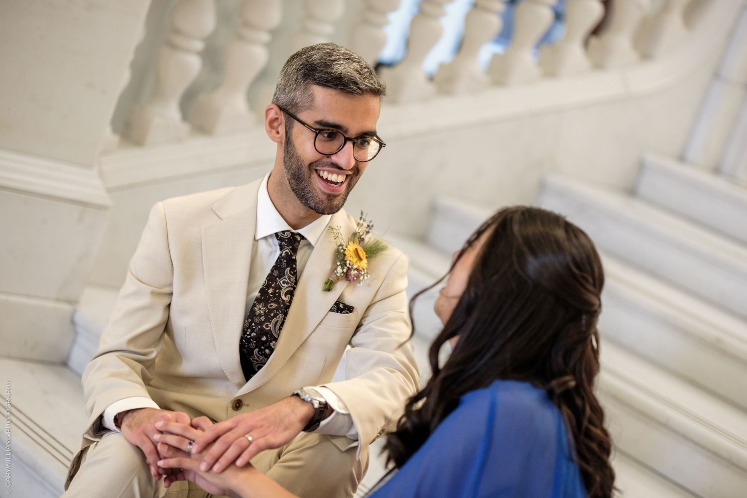 Groom in beige suit with floral tie smiles at bride during wedding day portraits on elegant staircase.