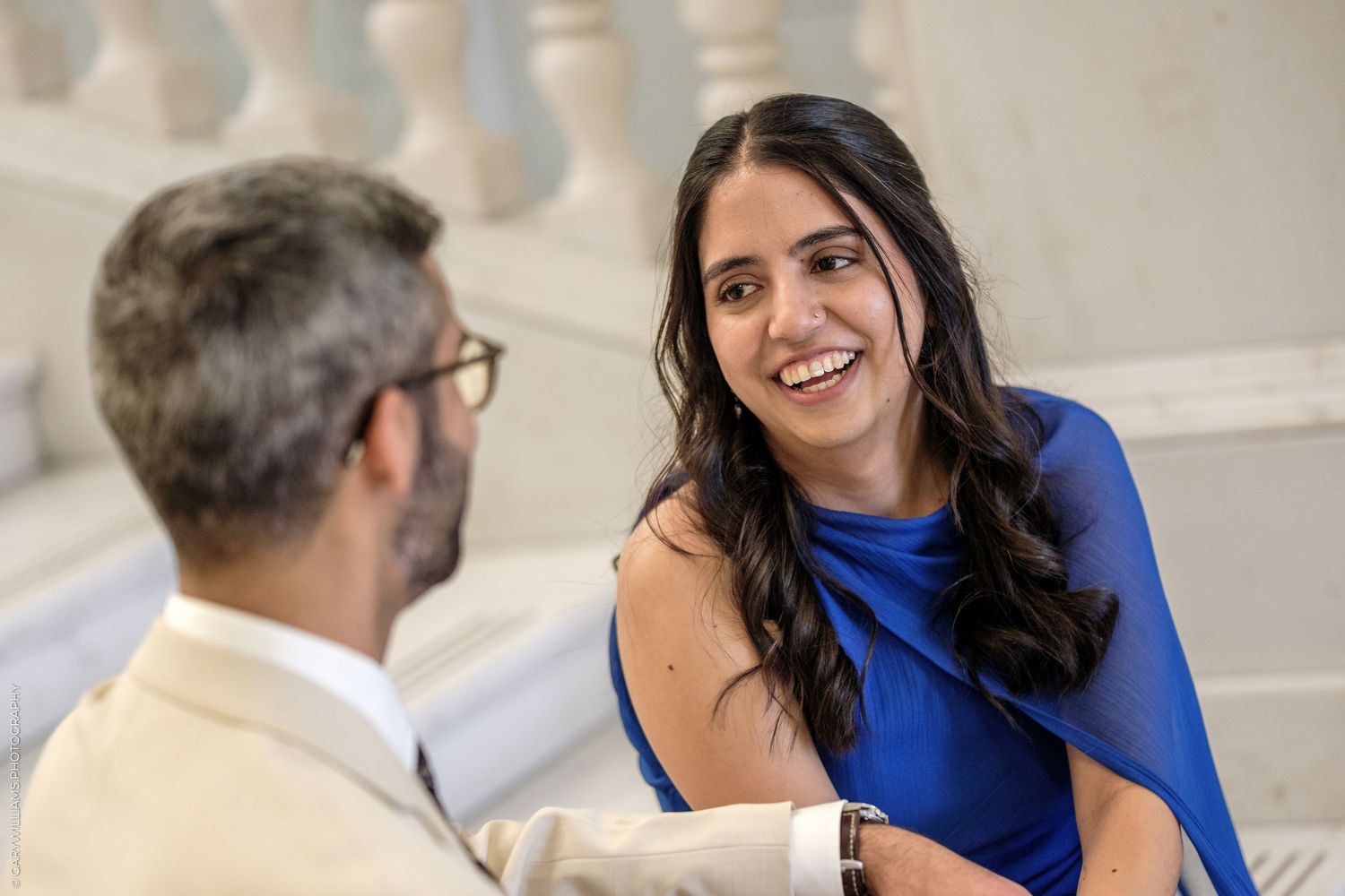A smiling woman in a blue dress engages in conversation with a man in a beige suit on elegant stairs.