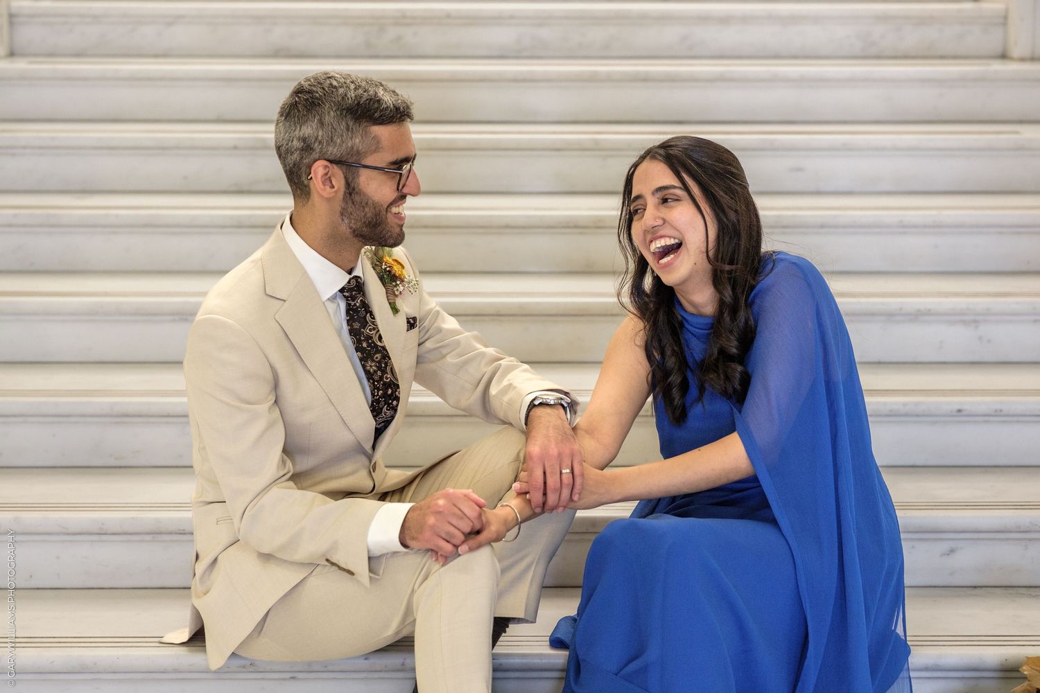 Couple in formal attire laughing together on marble steps, man in tan suit and woman in blue dress.