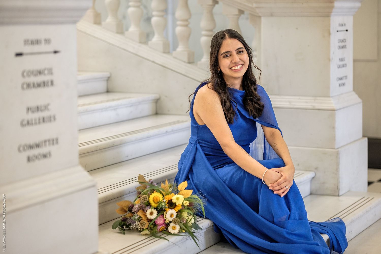 Woman in blue gown sitting on marble steps with floral bouquet at city hall.