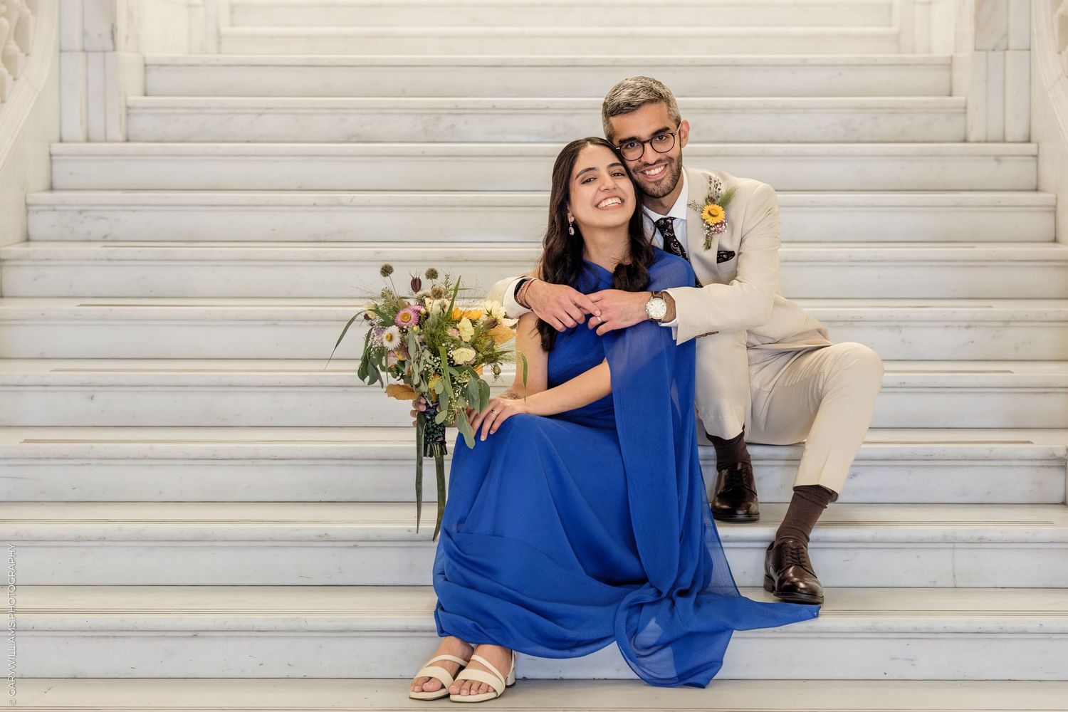 Couple in blue dress and tan suit sitting on marble steps with wildflower bouquet, elegant wedding portrait.