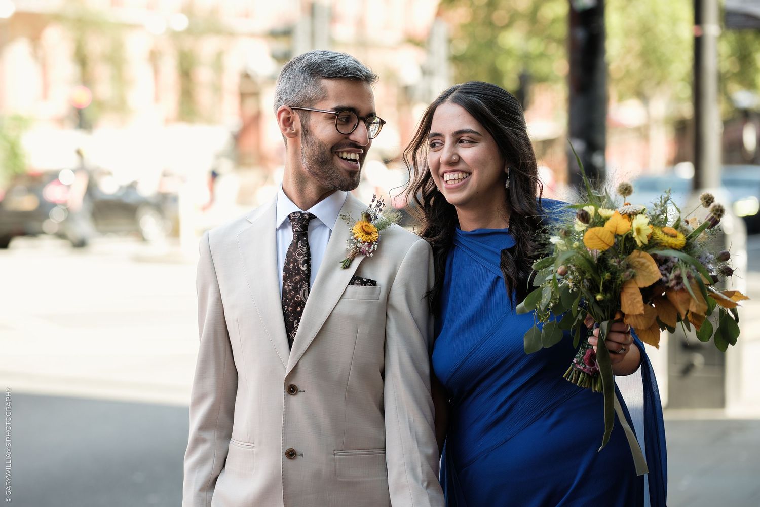 Happy couple in wedding attire, man in beige suit and woman in blue dress holding yellow flower bouquet outdoors.