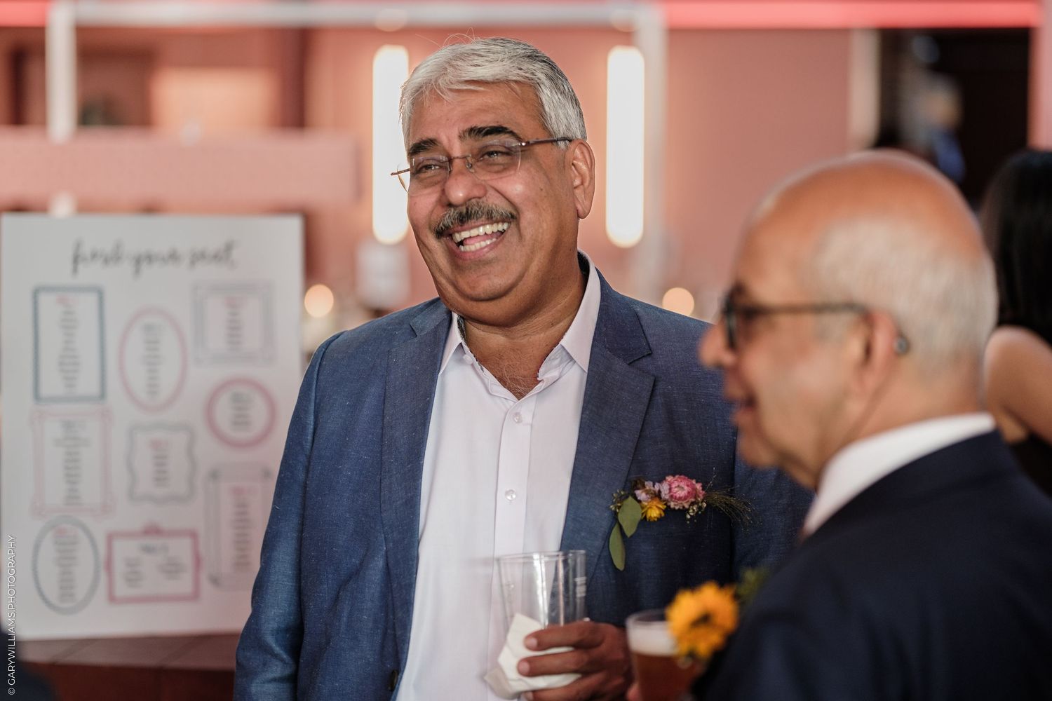 A man in a blue blazer smiles and holds a drink at an elegant indoor event.