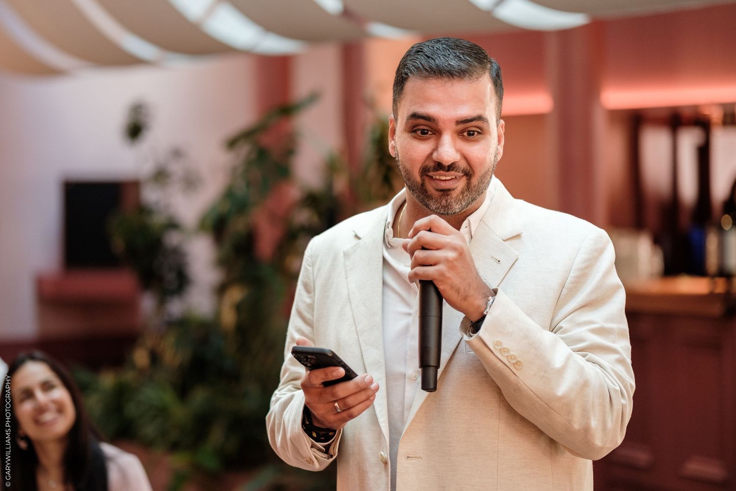 Man in white blazer smiling, holding microphone and phone, speaking at an indoor event.