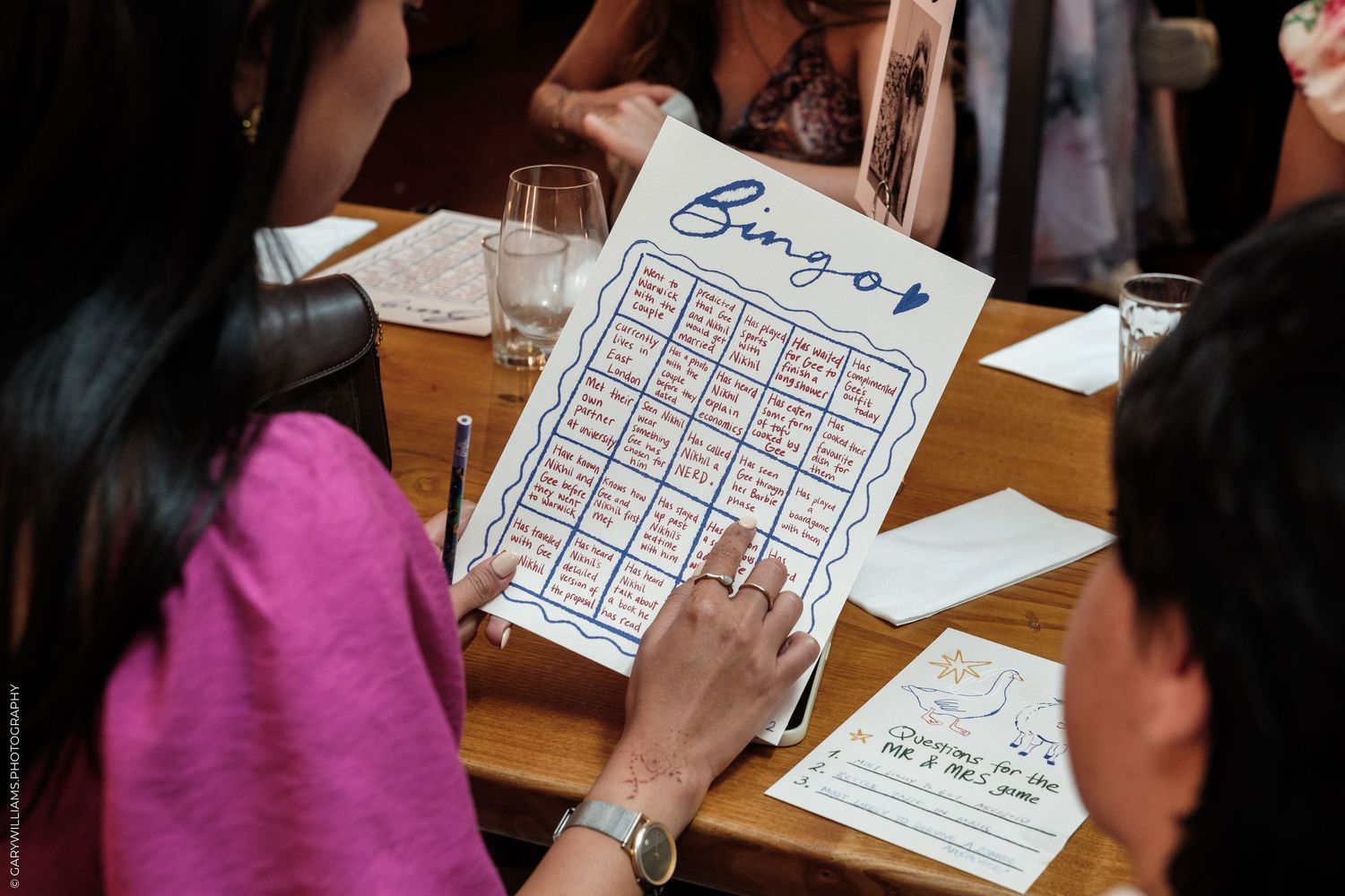 People playing wedding bingo game at a reception table, with a handmade bingo card visible.