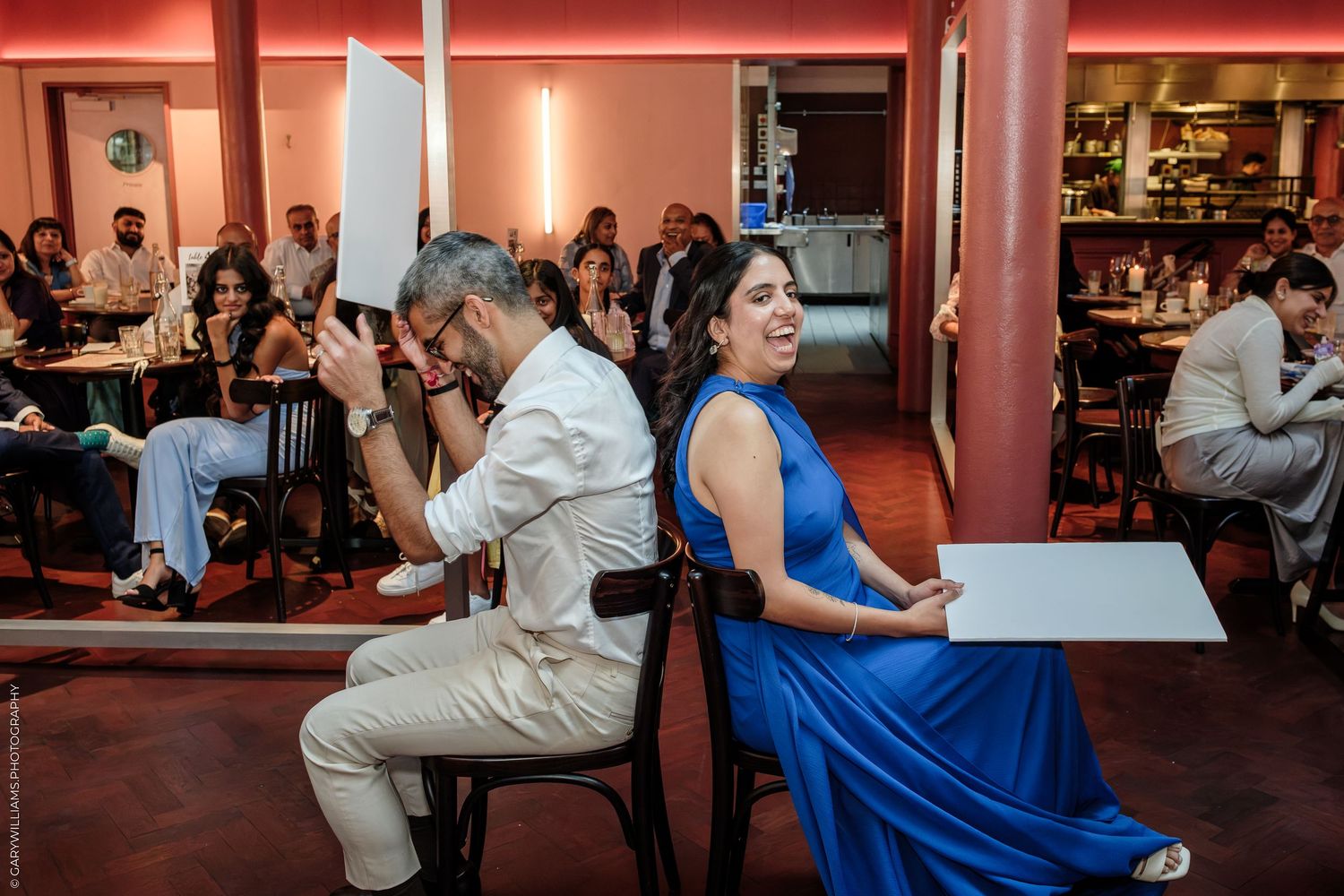 A man in white and woman in blue dress sit back-to-back at a restaurant event, smiling and holding white boards.