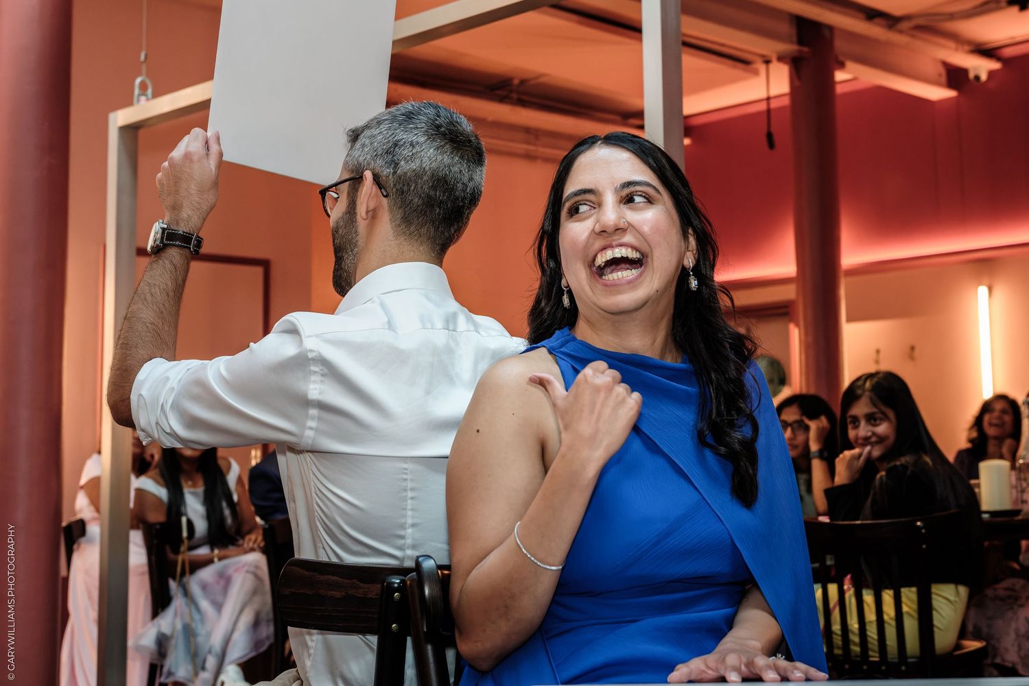 Woman in blue dress laughing joyfully at an elegant indoor event, with guests visible in the background.