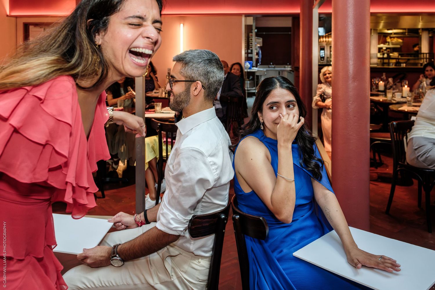 Three friends laughing and sharing a joyful moment at a restaurant, woman in red dress, man in white shirt, woman in blue dress.