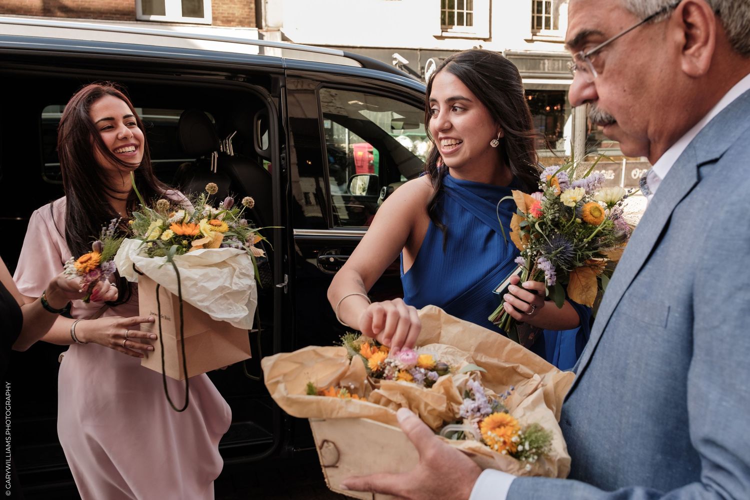 Two women and a man holding colorful flower bouquets outdoors near a black vehicle on a city street.