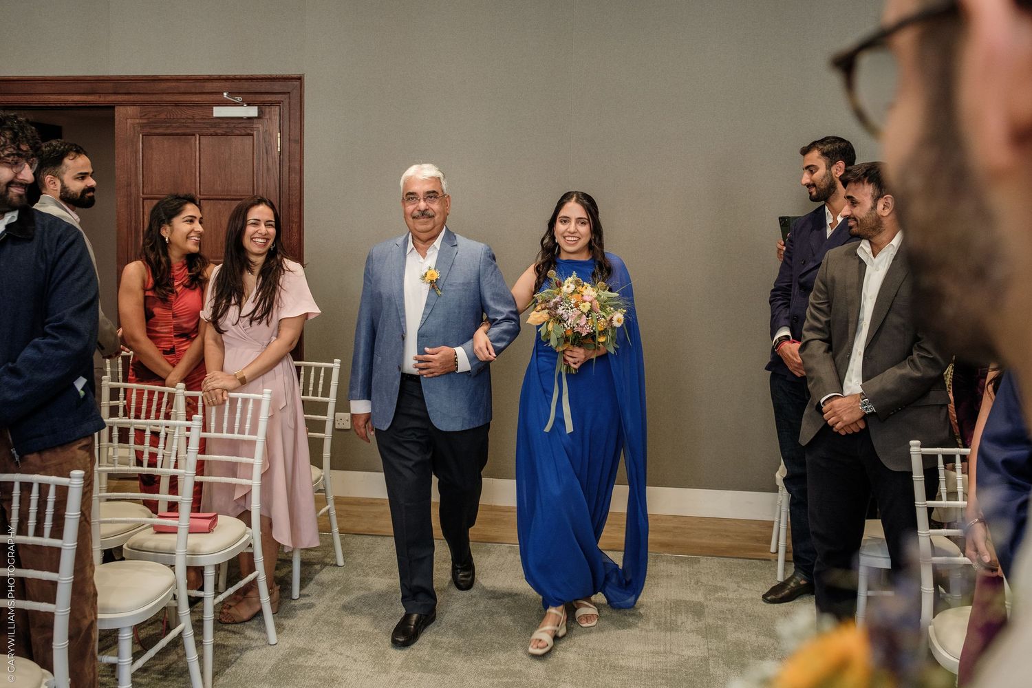Bride in blue gown walks down aisle with escort at elegant indoor wedding ceremony.