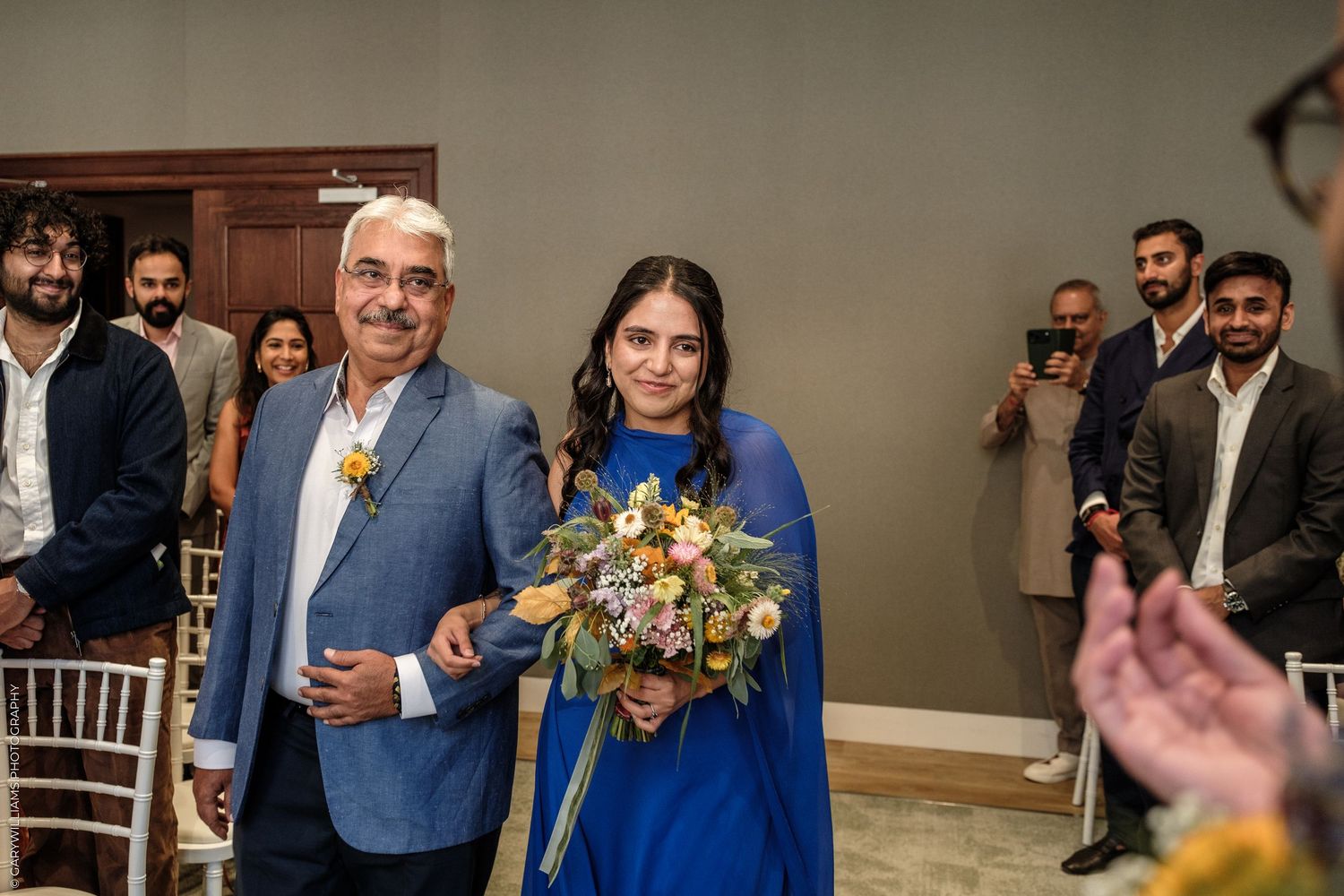 Woman in blue dress holding bouquet walks down aisle with older man in blue suit at indoor wedding ceremony.