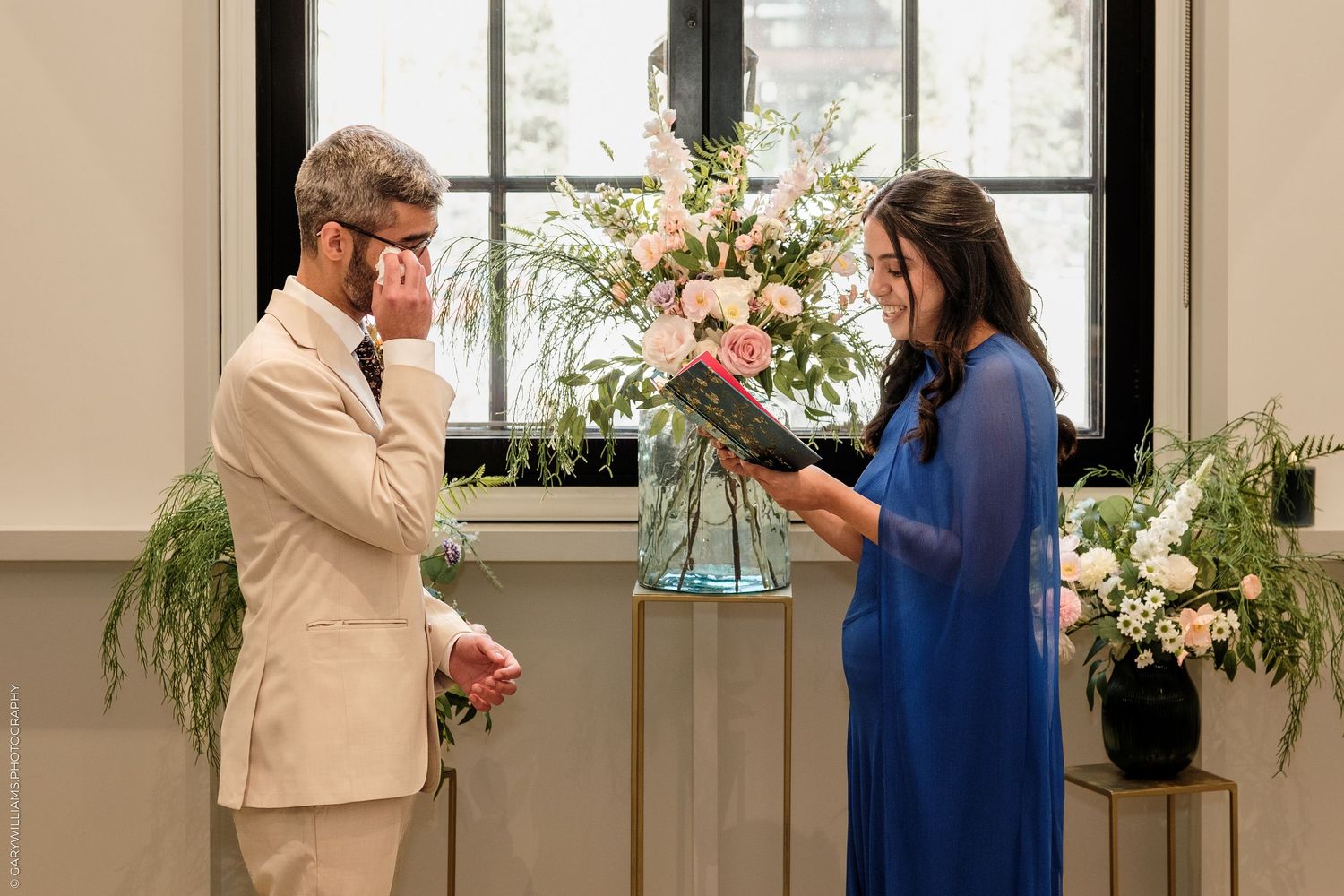 Emotional groom wiping tears as bride reads vows at elegant indoor wedding ceremony with floral arrangements.