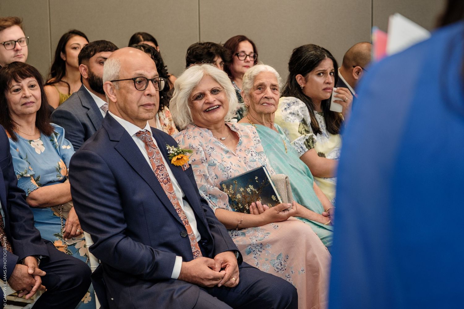 Elegantly dressed guests smile and watch attentively during a formal multicultural celebration or wedding ceremony indoors.