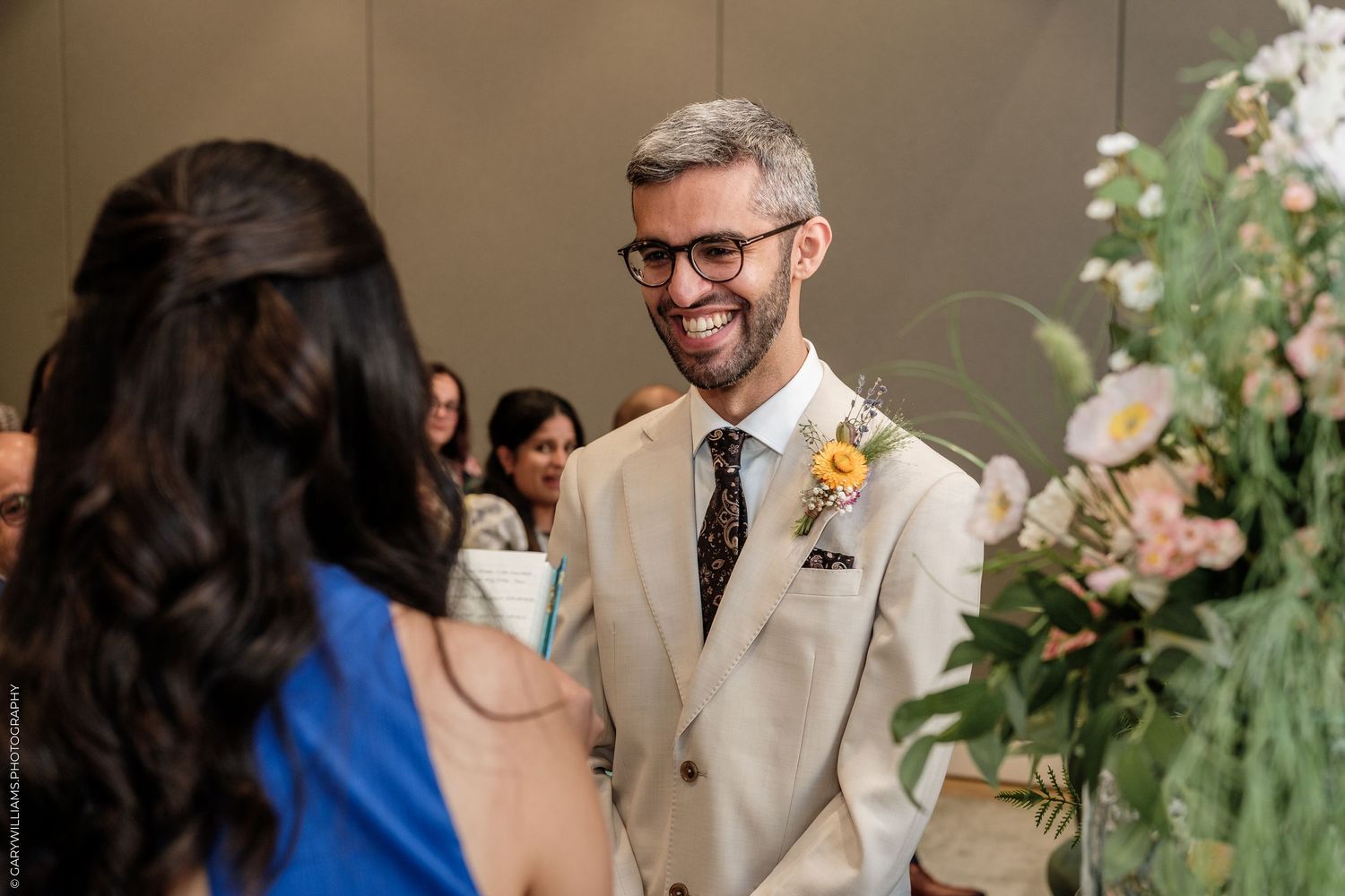 Groom in beige suit with sunflower boutonniere smiling at bride during wedding ceremony.