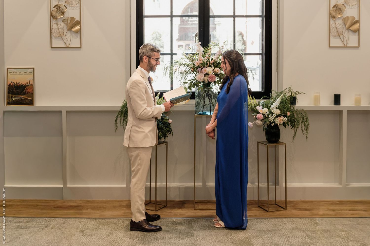 Couple exchanging vows at elegant indoor wedding ceremony with floral arrangements and large windows.