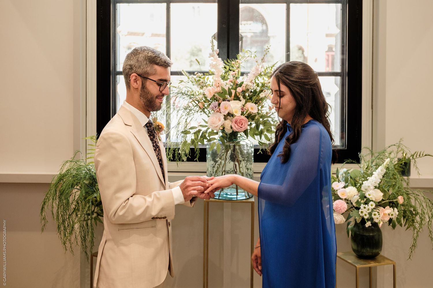 Groom in beige suit places ring on bride in blue dress during intimate wedding ceremony with floral arrangements.