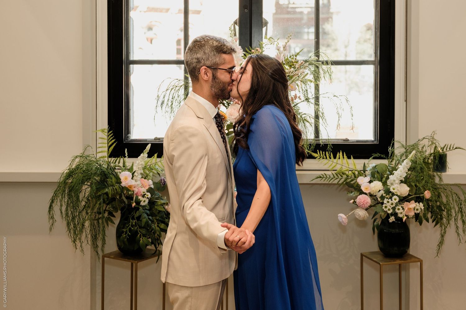 Couple in blue and beige sharing a kiss at an elegant indoor wedding ceremony.