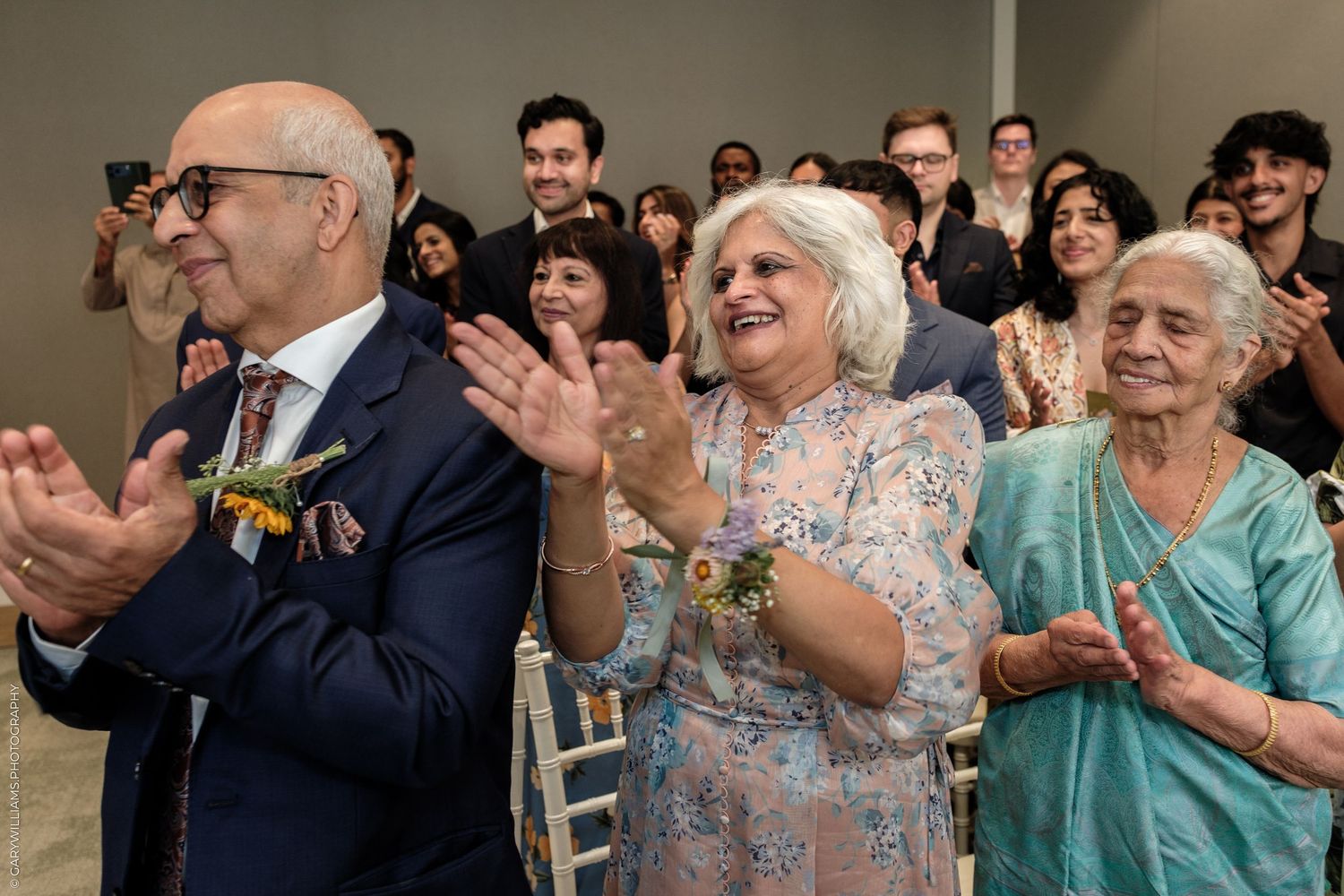 Joyful guests clapping and smiling at a wedding ceremony, dressed in elegant formal and traditional attire.