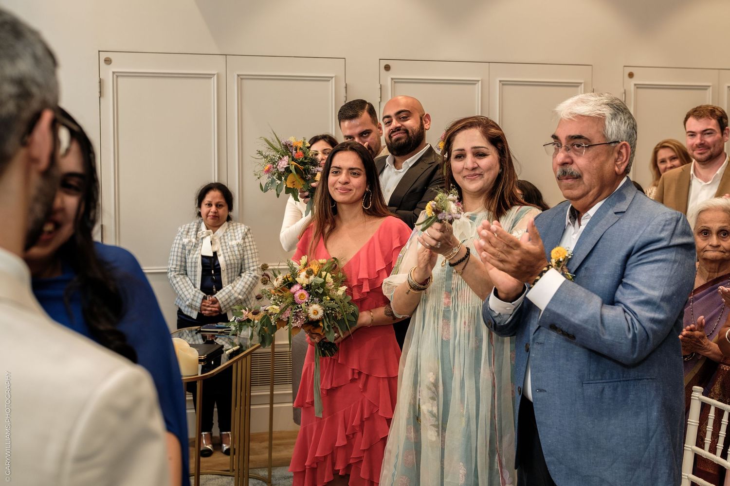 Guests clapping and smiling at an indoor wedding ceremony, with bridesmaids holding floral bouquets in a bright venue.