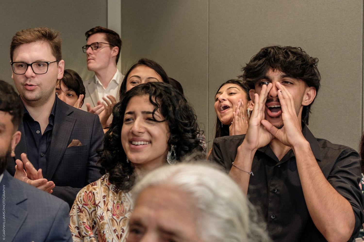 Excited crowd at an event, with a young man cupping his hands around his mouth to cheer loudly.