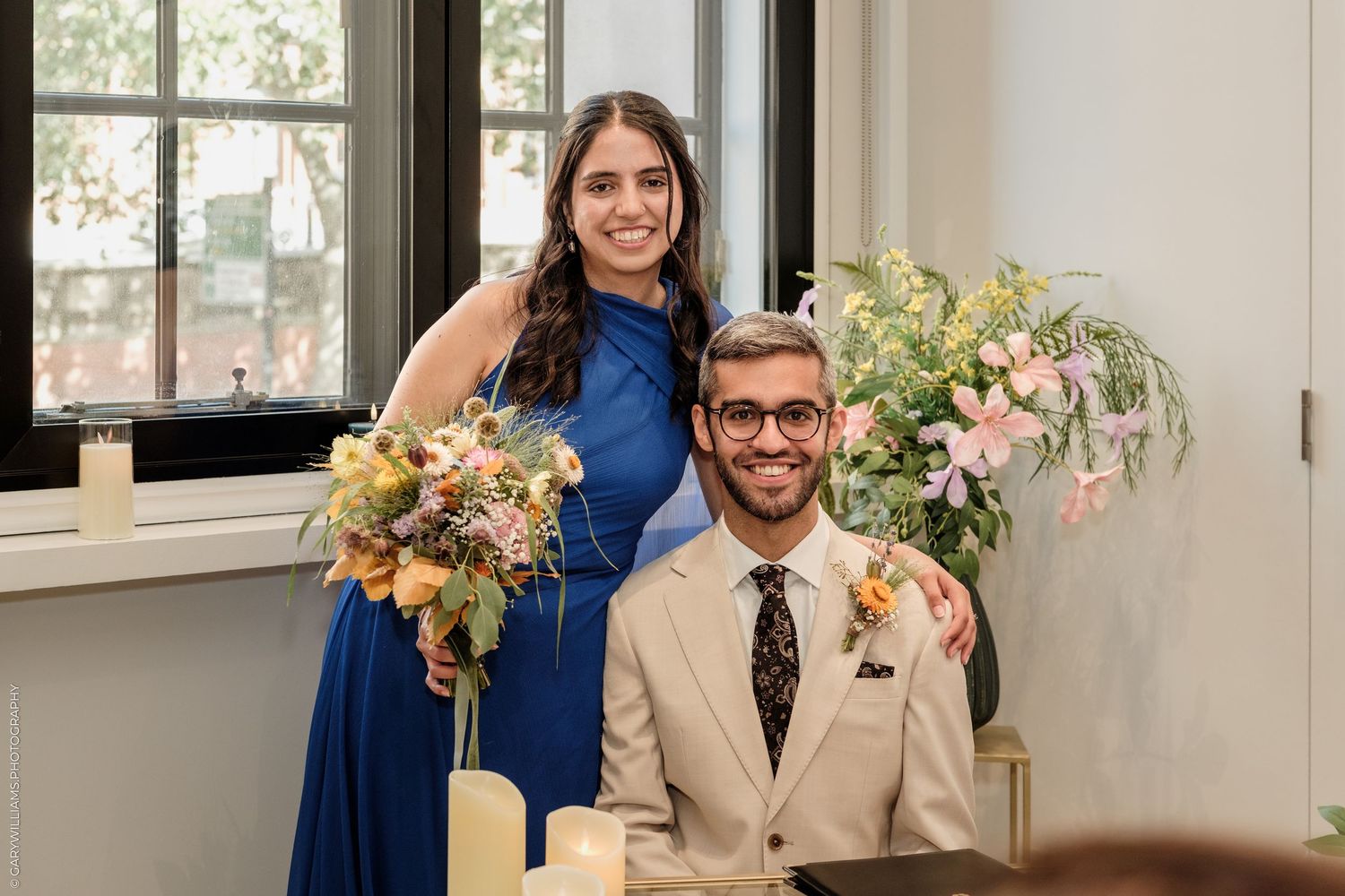 Bride in blue dress holding floral bouquet stands behind groom in beige suit at elegant wedding reception.