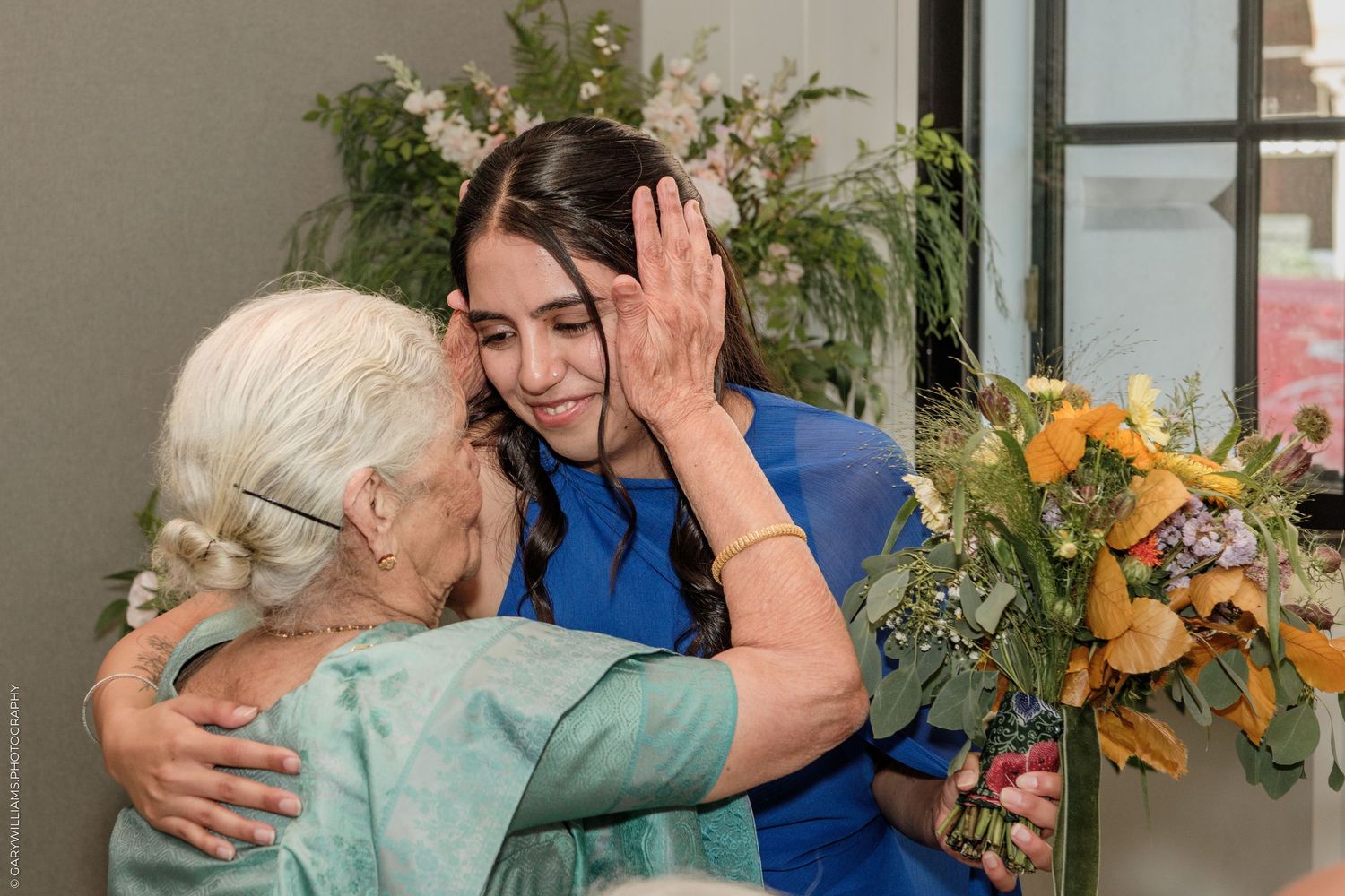 Two women share a warm embrace, one elderly in teal, the other younger in blue holding colorful flowers.