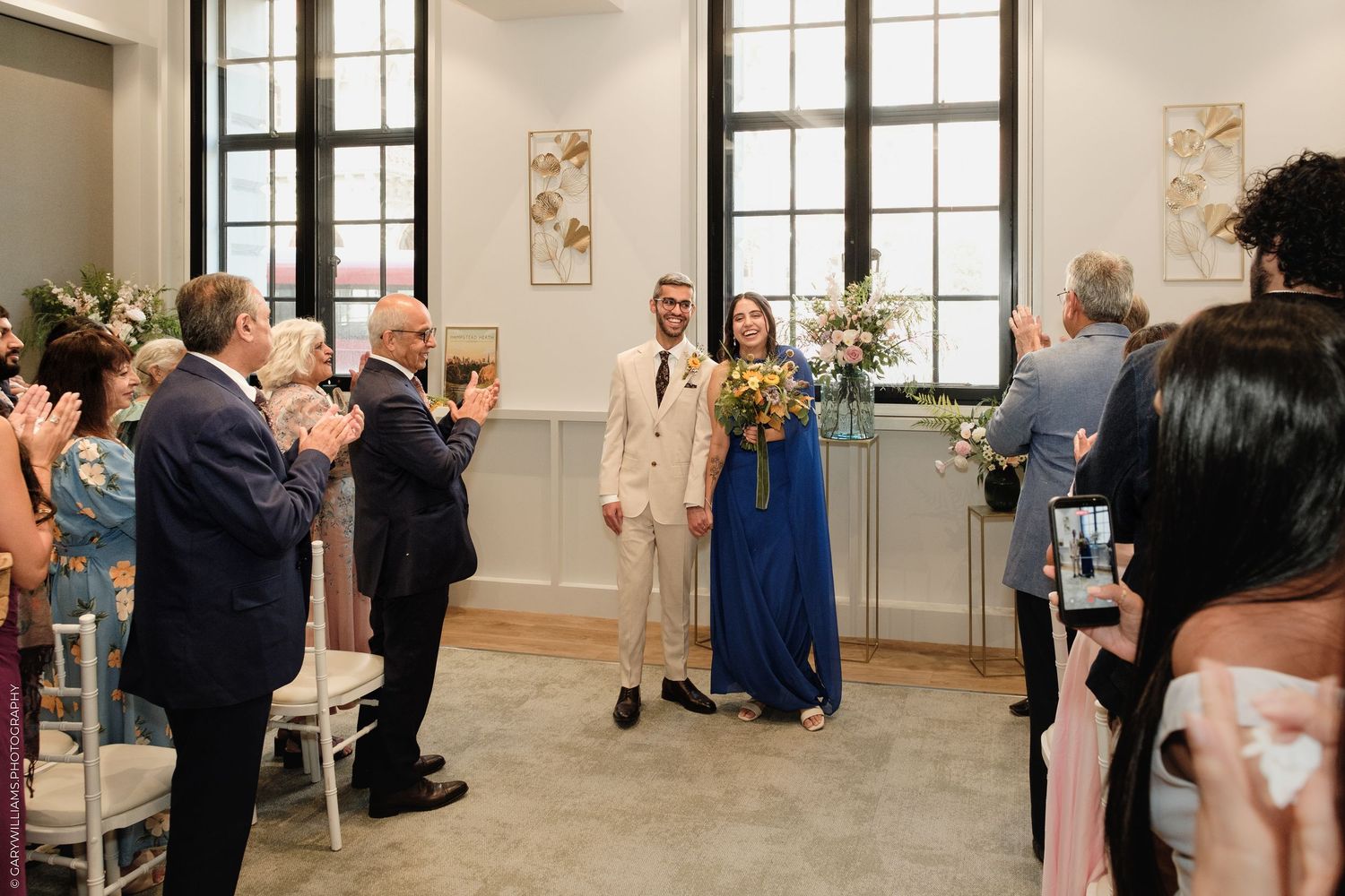 Bride and groom walk down the aisle as guests applaud in an elegant wedding venue with large windows.