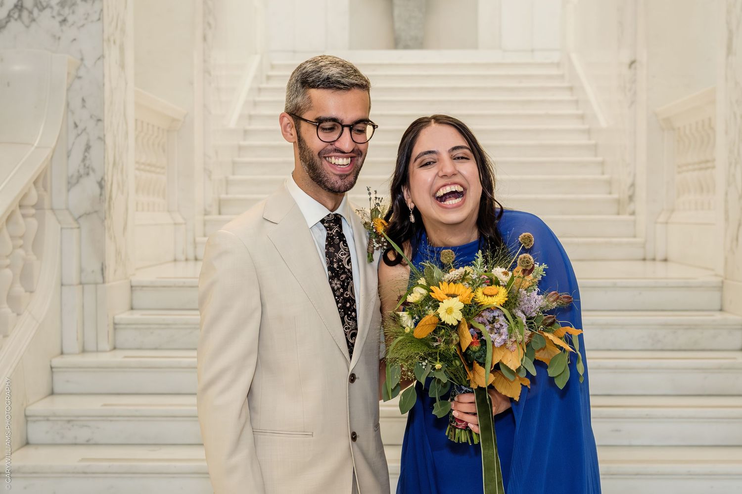 Happy couple posing on marble staircase, man in beige suit, woman in blue dress holding colorful bouquet at wedding.
