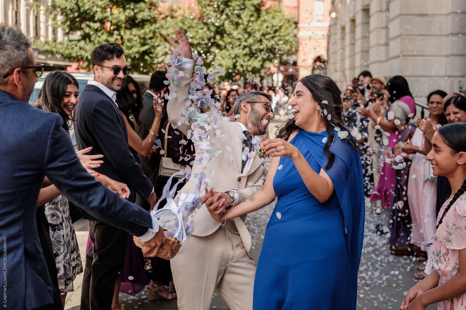Joyful wedding couple walks through confetti shower surrounded by celebrating guests outdoors.