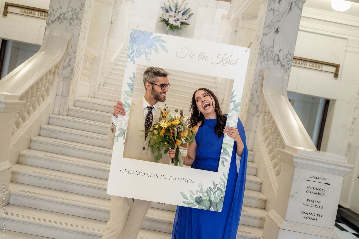 Couple holds decorative wedding photo frame on grand staircase, woman in blue dress holds yellow flowers.