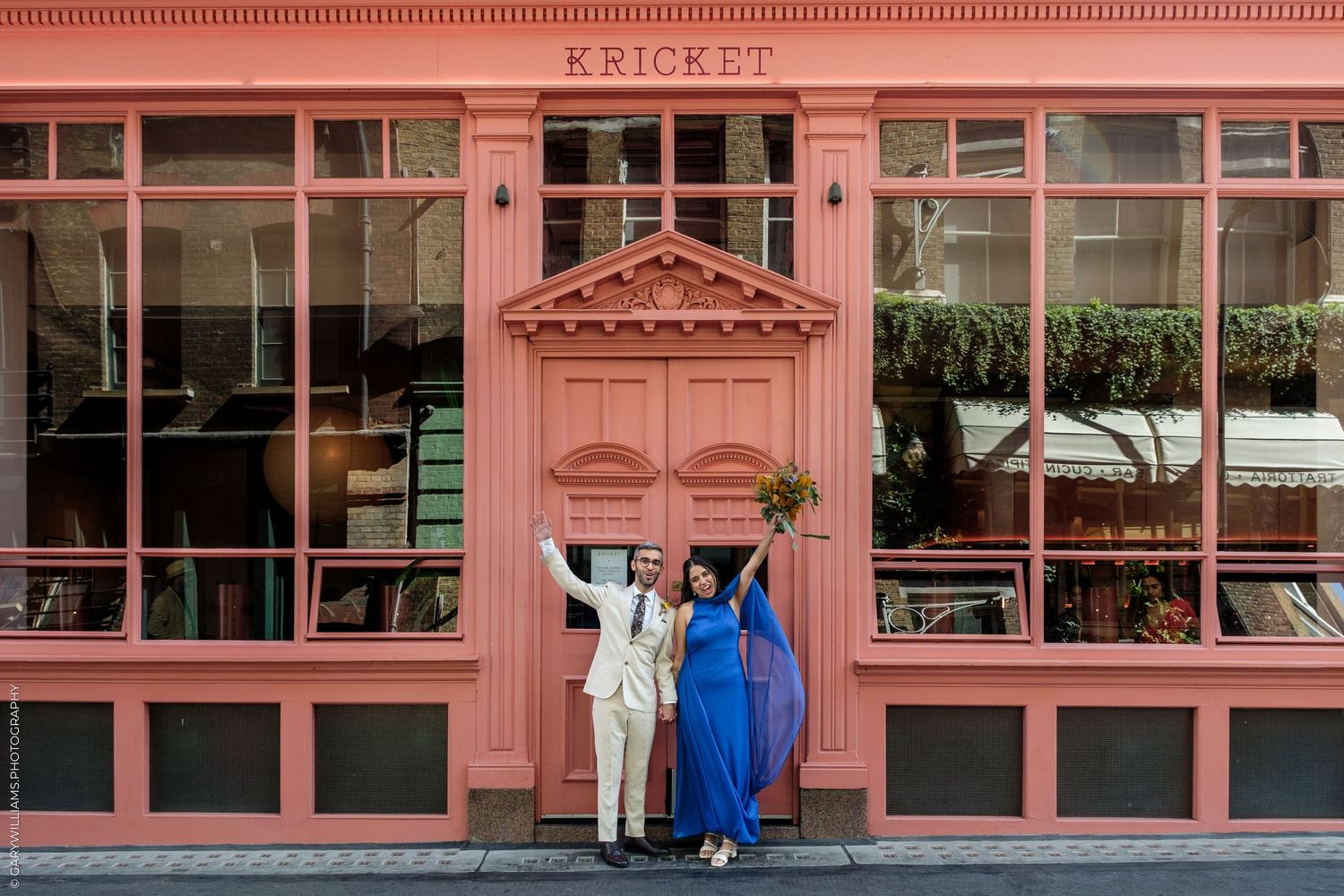 Couple celebrating outside Kricket restaurant, woman in blue gown and man in white suit, pink facade backdrop.