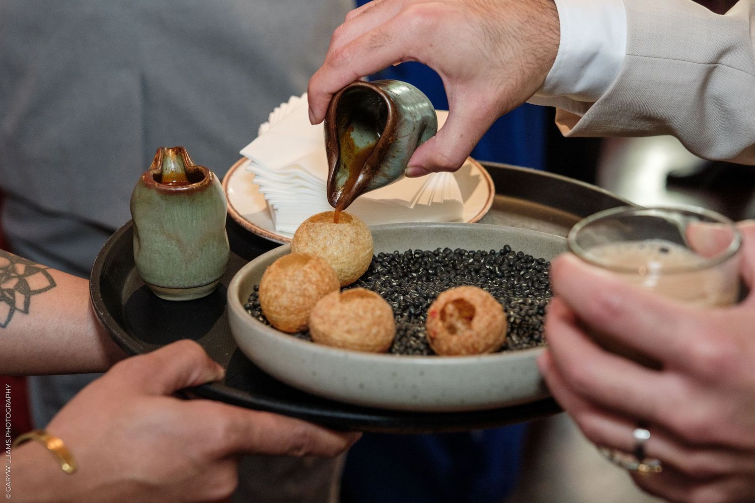 Chef pouring sauce over golden fried dough balls in a dark bowl filled with black seeds at a fine dining event.