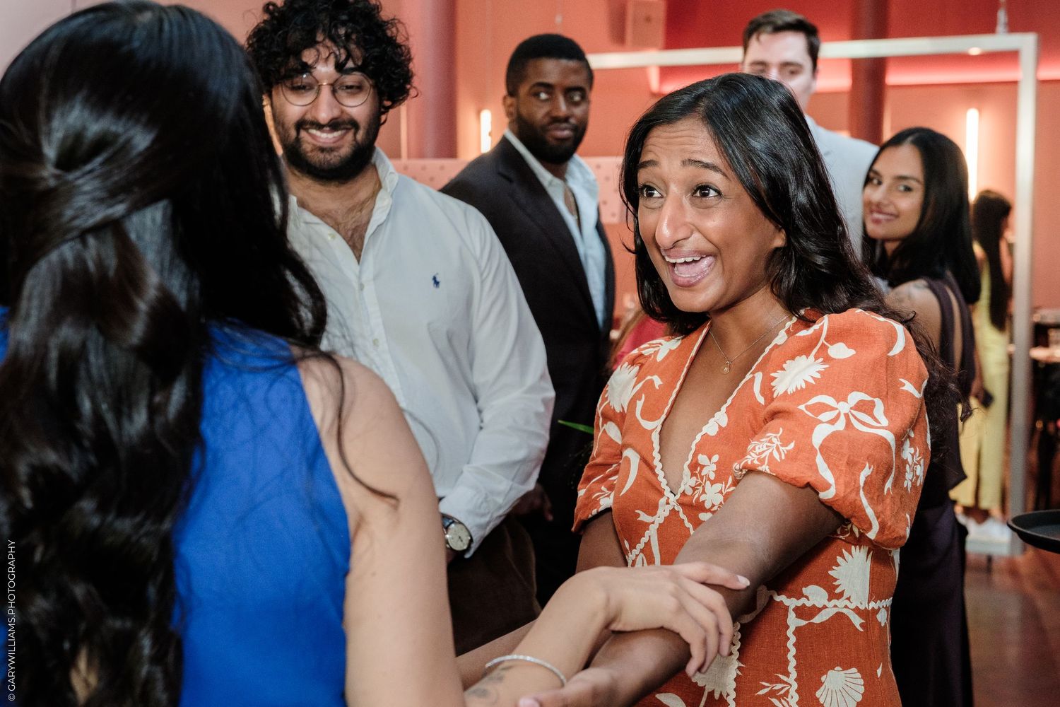 Group of young professionals socializing at an indoor event, woman in orange floral dress smiling warmly.
