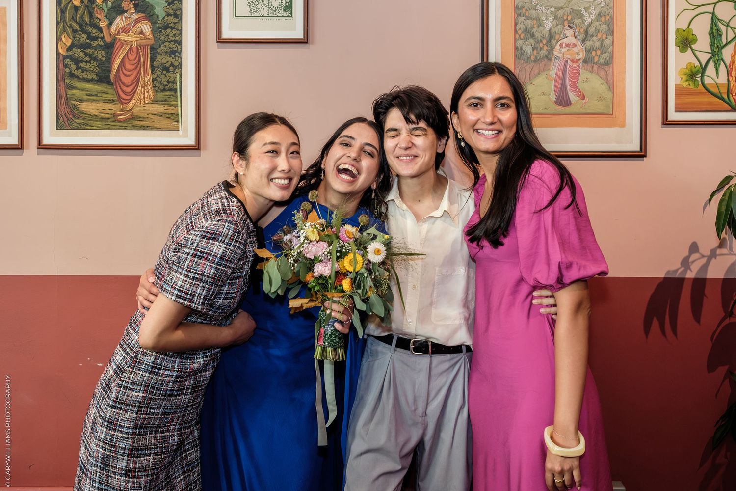 Four women smiling and hugging, one holding a colorful bouquet, posing together in a warmly decorated room.