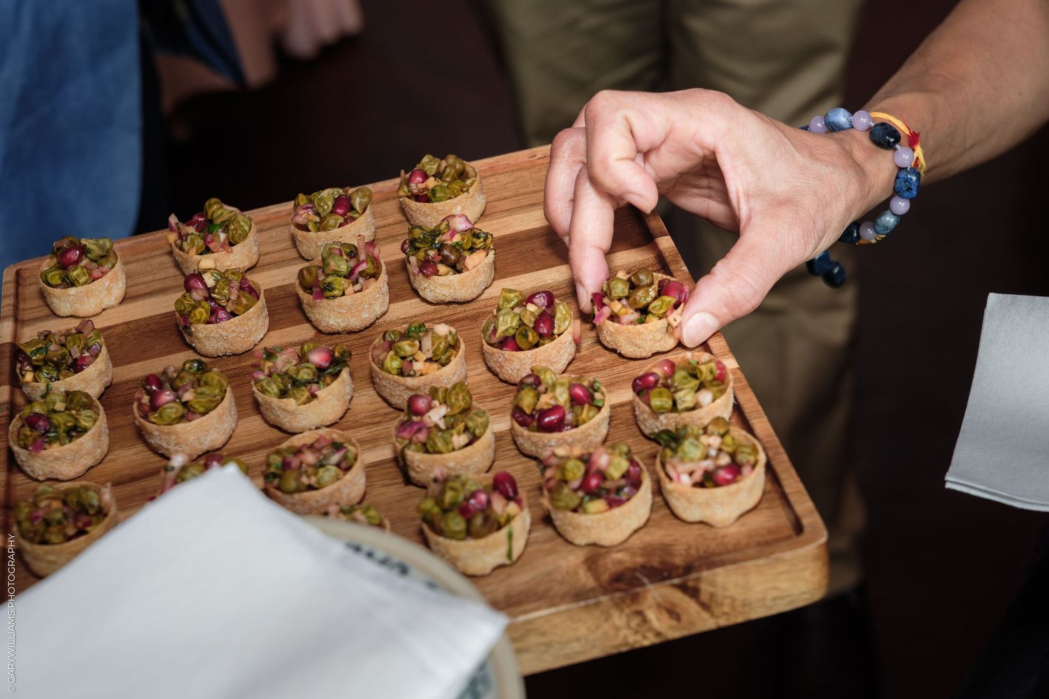 Hand selecting a gourmet appetizer from a wooden tray of mini tartlets topped with colorful garnishes at a catered event.