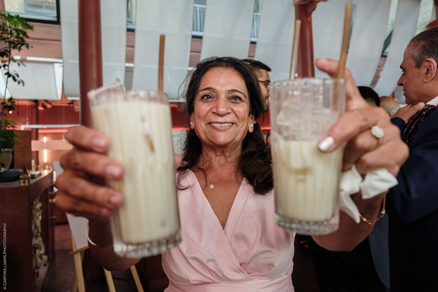Woman in pink dress smiling and holding two milky cocktails with straws at a festive outdoor gathering.