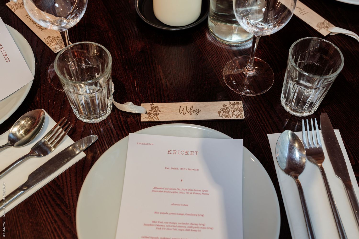 Elegant restaurant place setting with menu, crystal glasses, cutlery, and a personalized name card on a dark wood table.