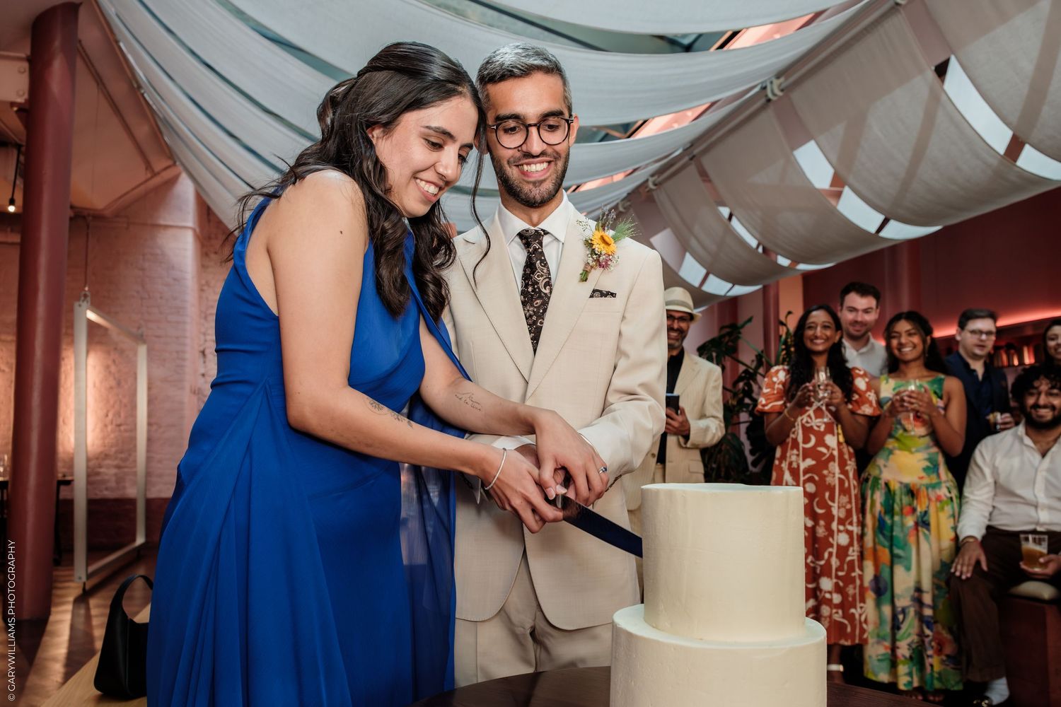 Bride in blue dress and groom in tan suit cutting white wedding cake together at elegant reception.