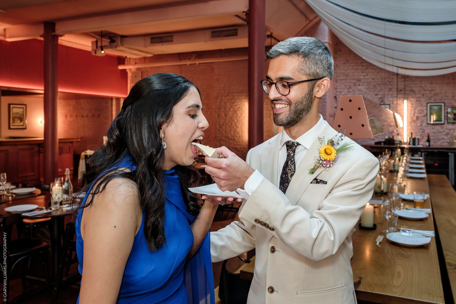 Groom in cream suit feeds wedding cake to bride in blue dress at elegant restaurant reception.