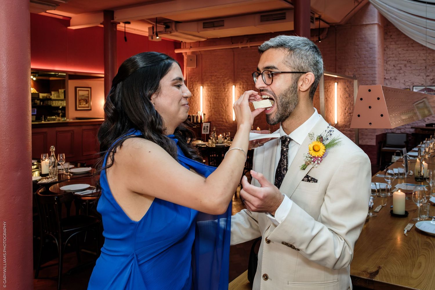 Bride in blue dress feeds groom in tan suit wedding cake at reception venue.