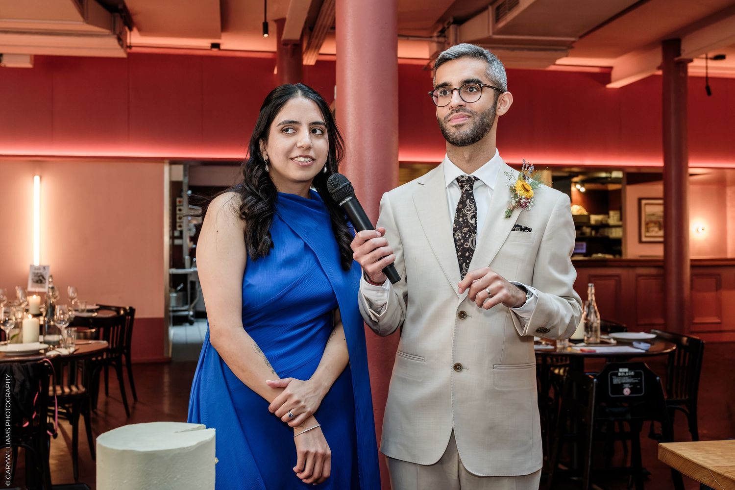 A woman in a blue dress and a man in a beige suit hold a microphone at an elegant wedding reception.