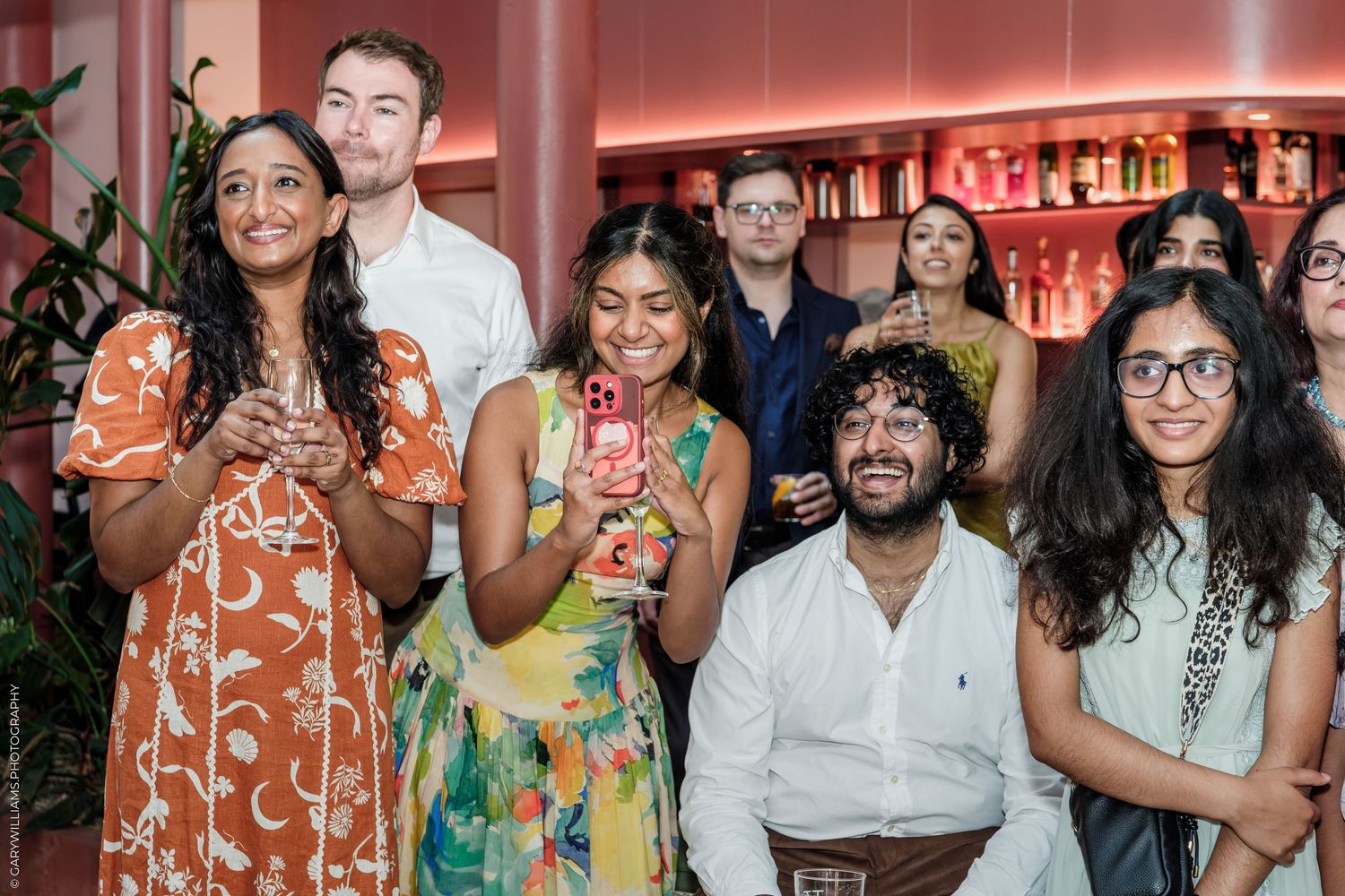 Diverse group of young adults smiling and celebrating at a social event in a stylish pink bar setting.