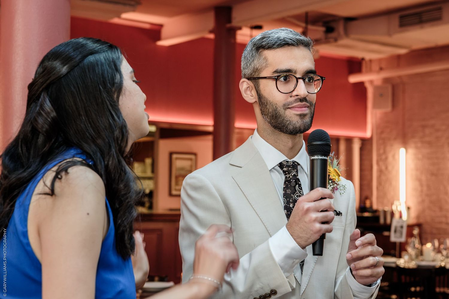Man in beige suit holding microphone speaks at event beside woman in blue dress in elegant venue.