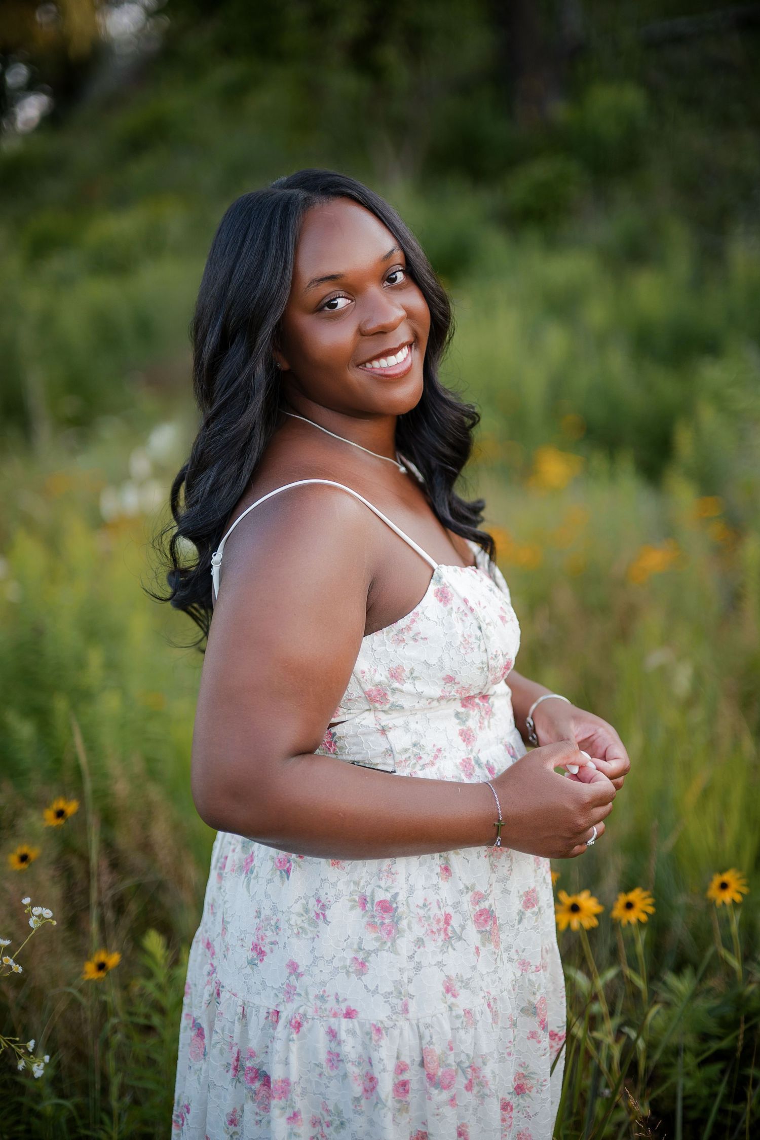 Young Black woman in floral sundress smiling outdoors among wildflowers and yellow blooms in a lush green field.