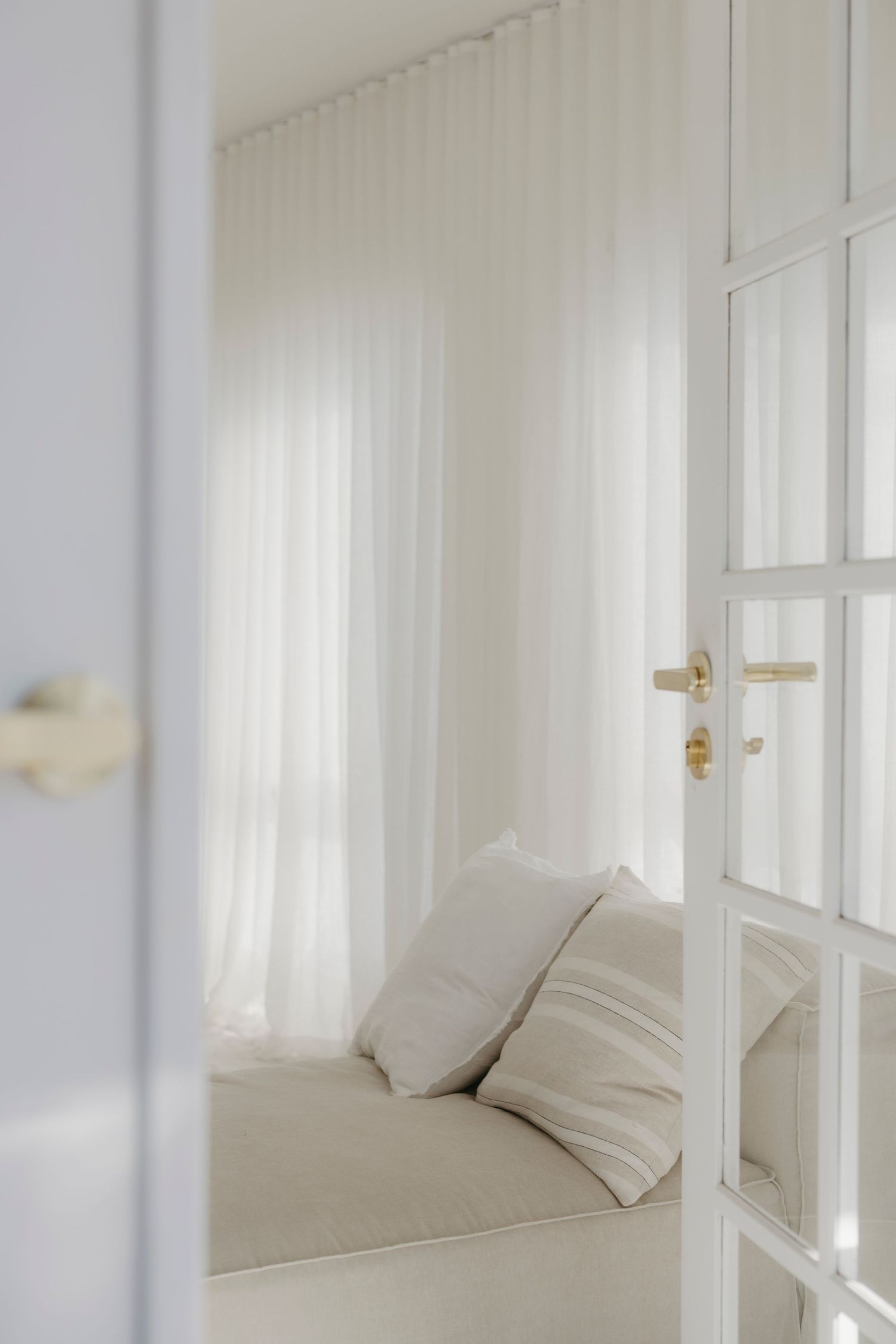 Minimalist white bedroom with sheer curtains, neutral cushions, and glass-paneled door.
