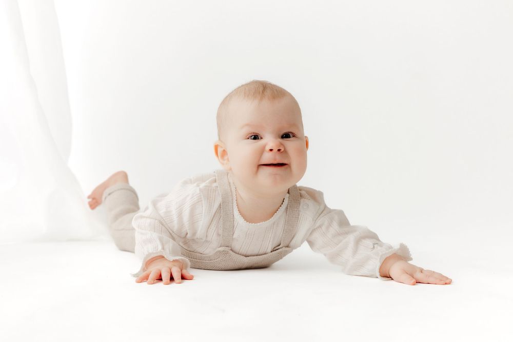 A happy baby in a white onesie crawls on a white floor, looking up with a bright smile.