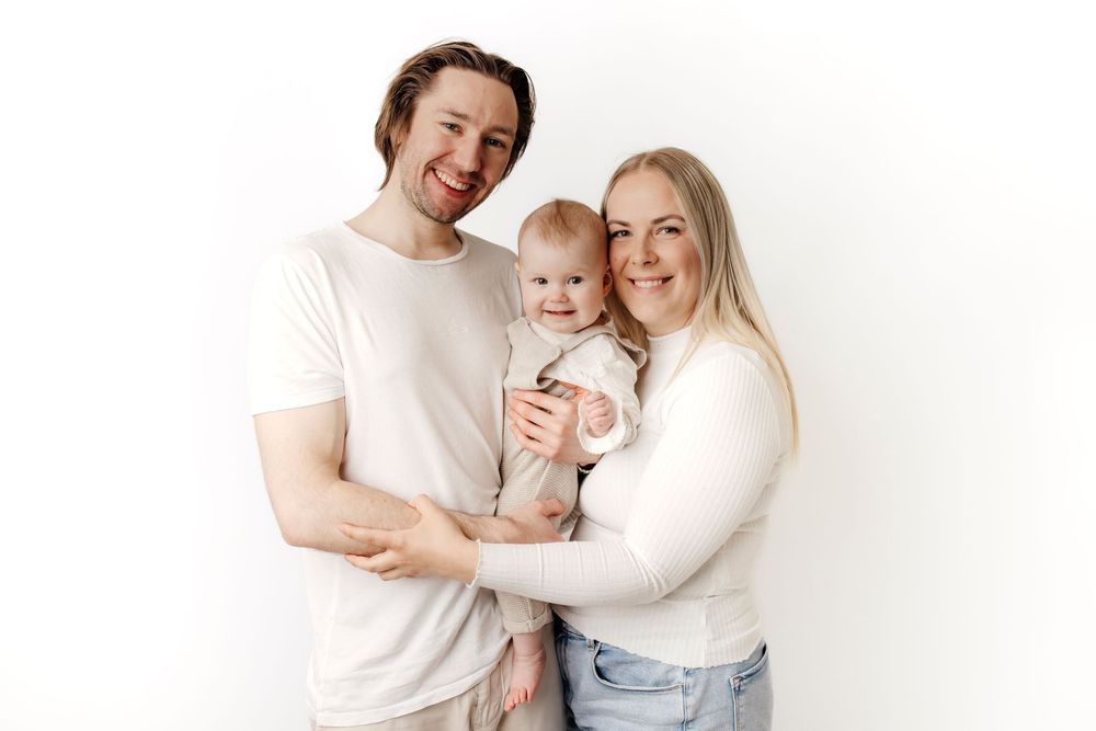 A happy young family with a man, woman, and baby posing together against a white background.