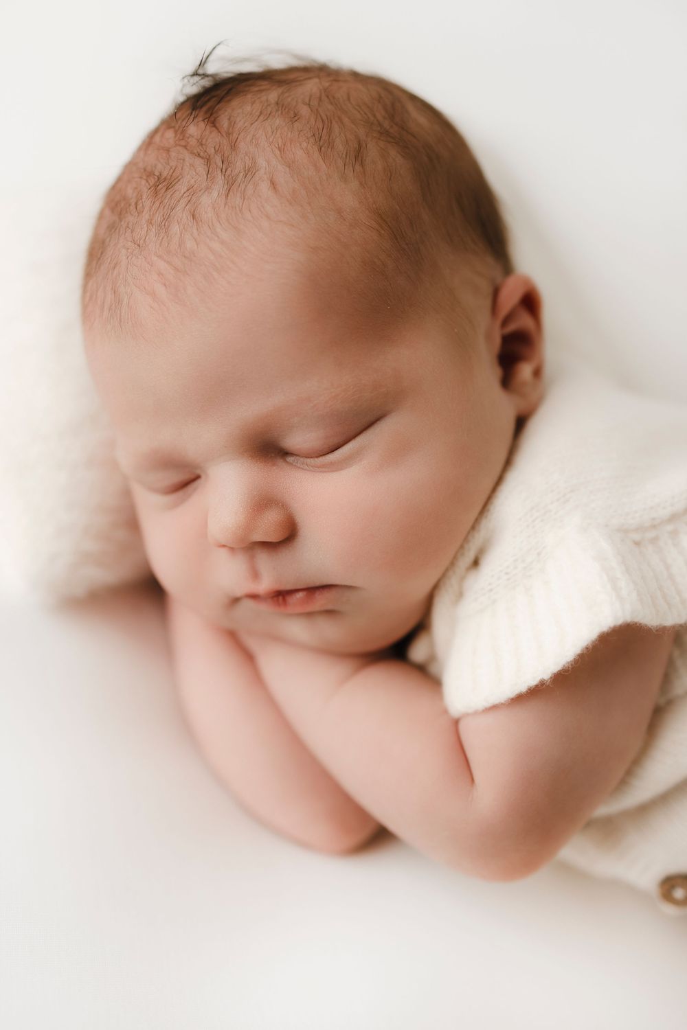 Peaceful newborn baby sleeping with hands tucked under chin, wearing a white knit romper on a soft white background.